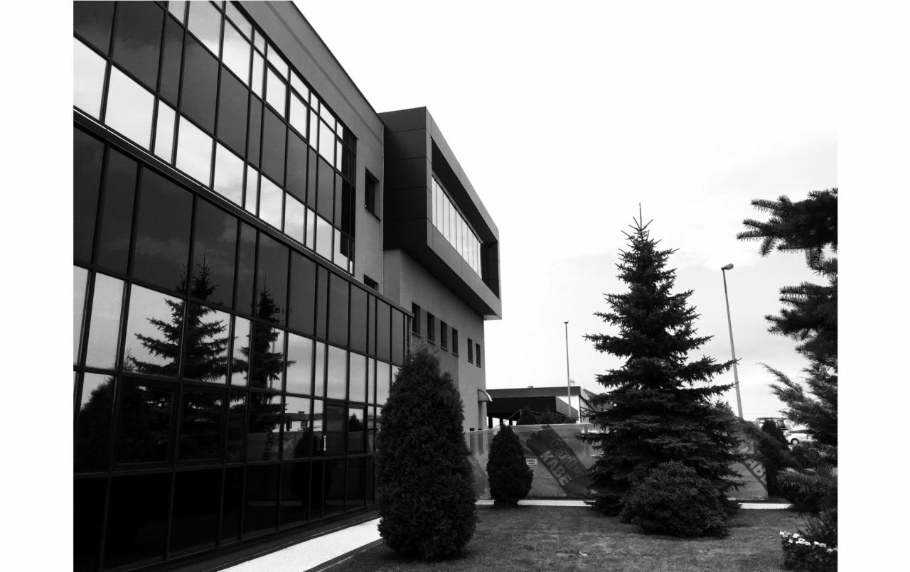 Czarno-białe zdjęcie nowoczesnego budynku usługowego z dużą ilością okien, w których odbijają się drzewa. Przed budynkiem rosną drzewa iglaste i trawa.
