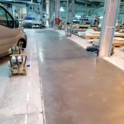 Świeżo wylana, szara posadzka w hali produkcyjnej, obok stoi szlifierka do betonu i zaparkowany samochód dostawczy.