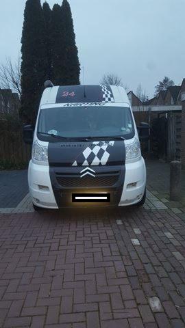 Biały bus z czarnymi elementami dekoracyjnymi w szachownicę na masce i dachu, zaparkowany na brukowanej nawierzchni przed posesją z zielonym drzewem w tle.