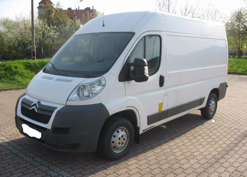 Biały bus Citroen Jumper na brukowanym parkingu, widok z boku, zarys zamku w tle.