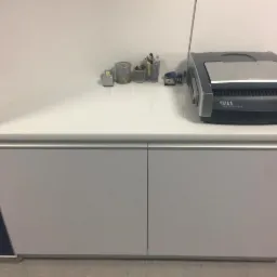 Biała szafka aktowa z frezowanym łukiem, na blacie biurowe akcesoria i bindownica, widok z bliska.