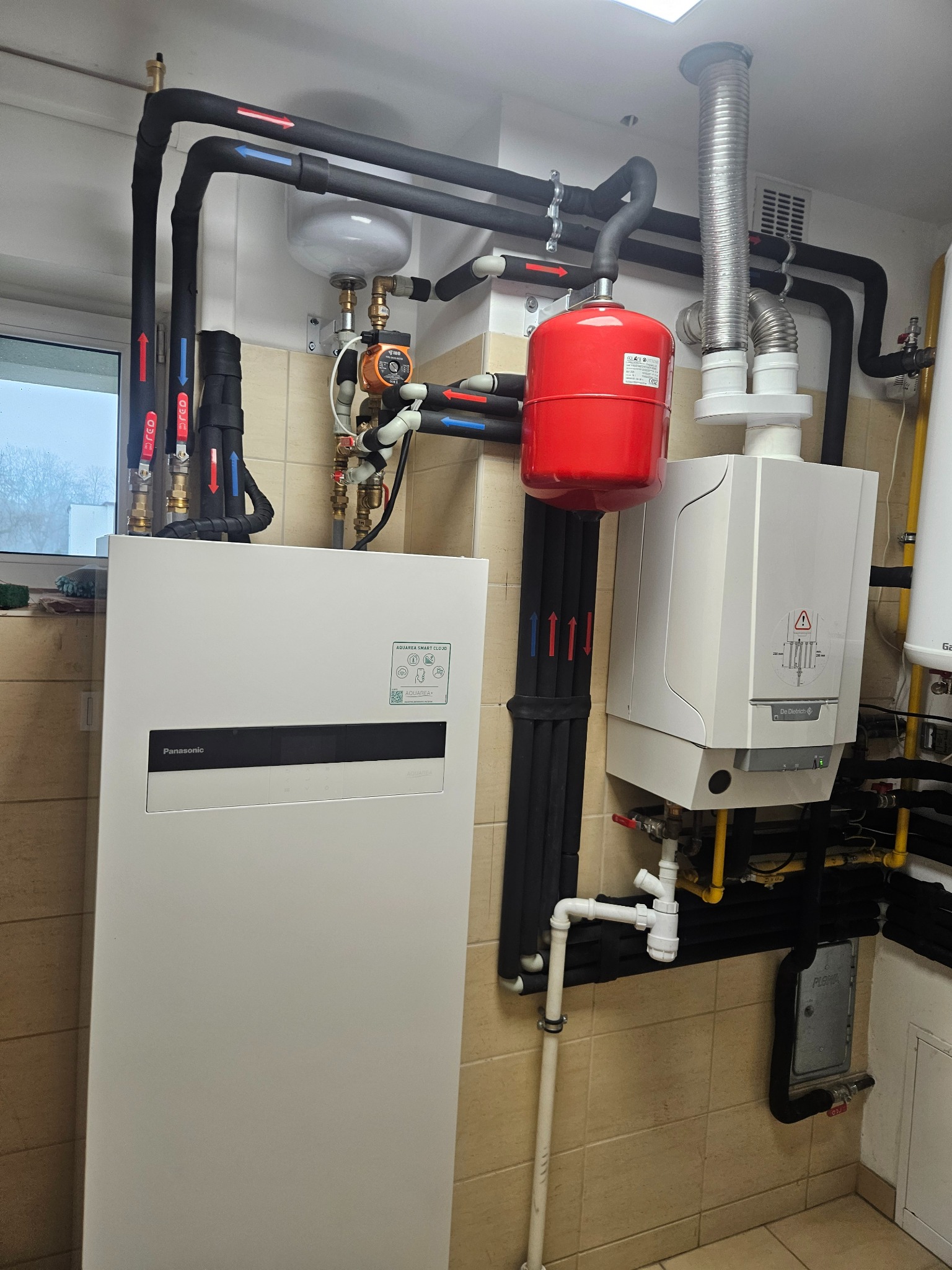 Pompa ciepła Panasonic All in One z instalacją, rurami w izolacji i zbiornikiem wyrównawczym w kotłowni z beżowymi płytkami. Widoczne okno.