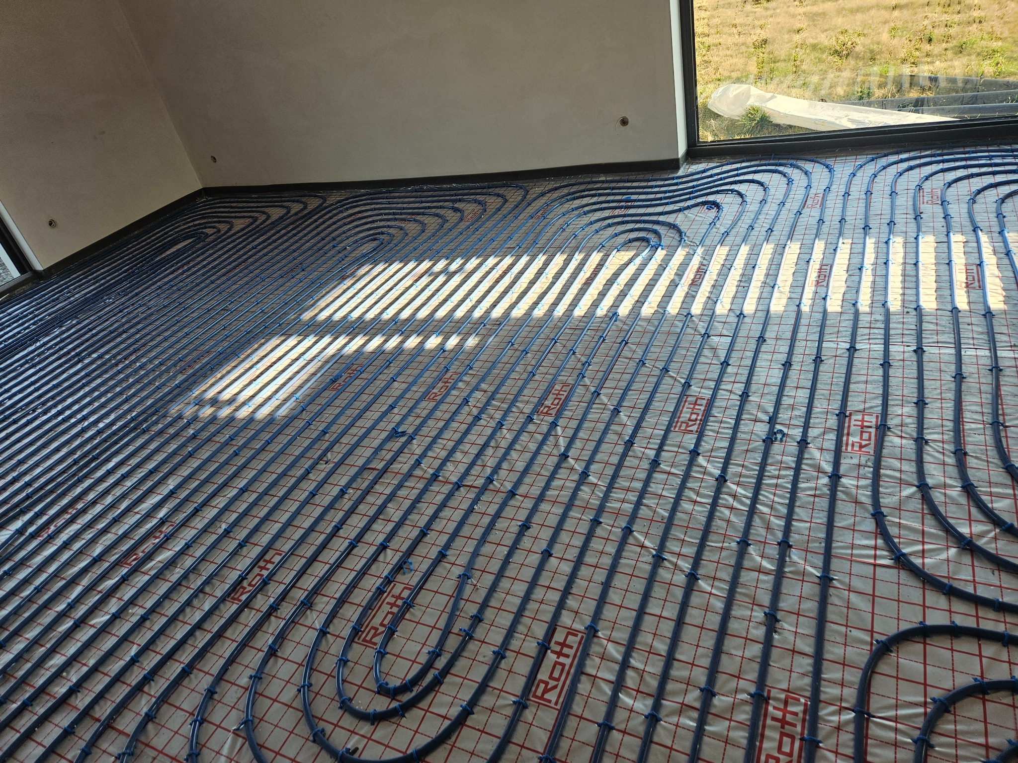 Ułożone niebieskie rury ogrzewania podłogowego marki Roth na folii z czerwoną kratką, widoczne okno i fragment poddasza. Promienie słoneczne padają na instalację.