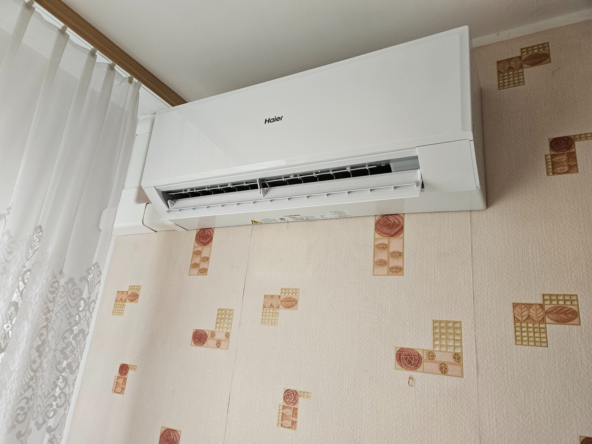 Biały klimatyzator Haier zamontowany na ścianie z wzorzystą tapetą, widoczny fragment zasłony. Urządzenie włączone, z otwartą klapką nawiewu.