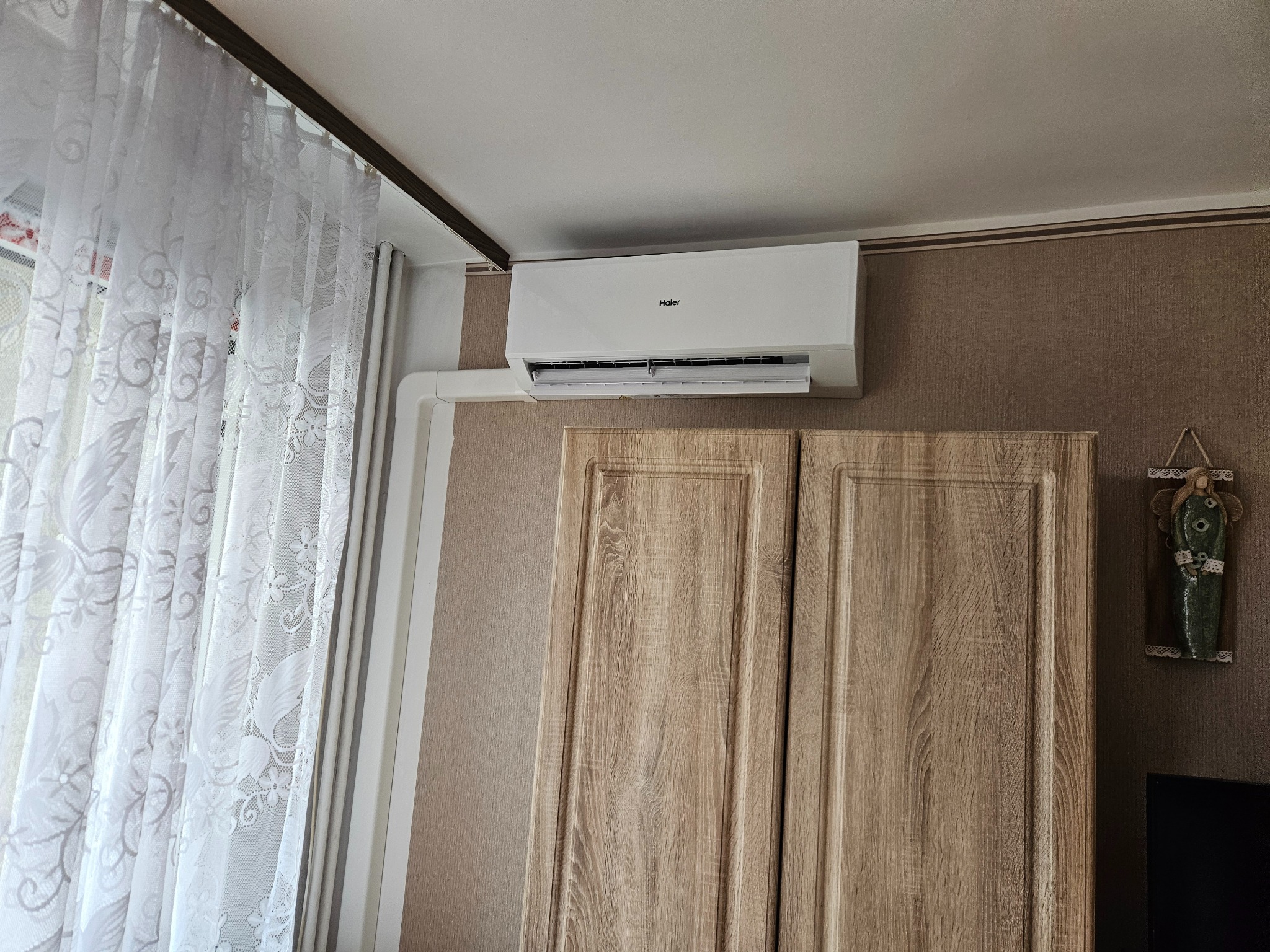 Biały klimatyzator Haier zamontowany nad drewnianą szafką, widoczne białe rury instalacyjne, zasłona w oknie. Ujęcie z dołu, wnętrze pokoju.
