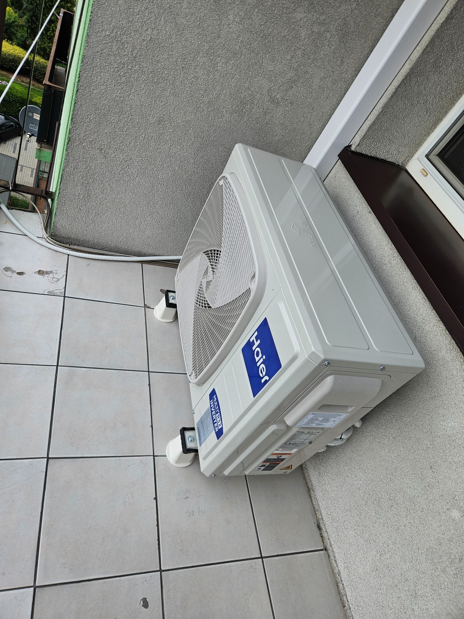 Zamontowana jednostka zewnętrzna klimatyzacji Haier na balkonie, widok z góry. Biała obudowa, szara ściana budynku, płytki podłogowe. Widoczne przewody i elementy montażowe.