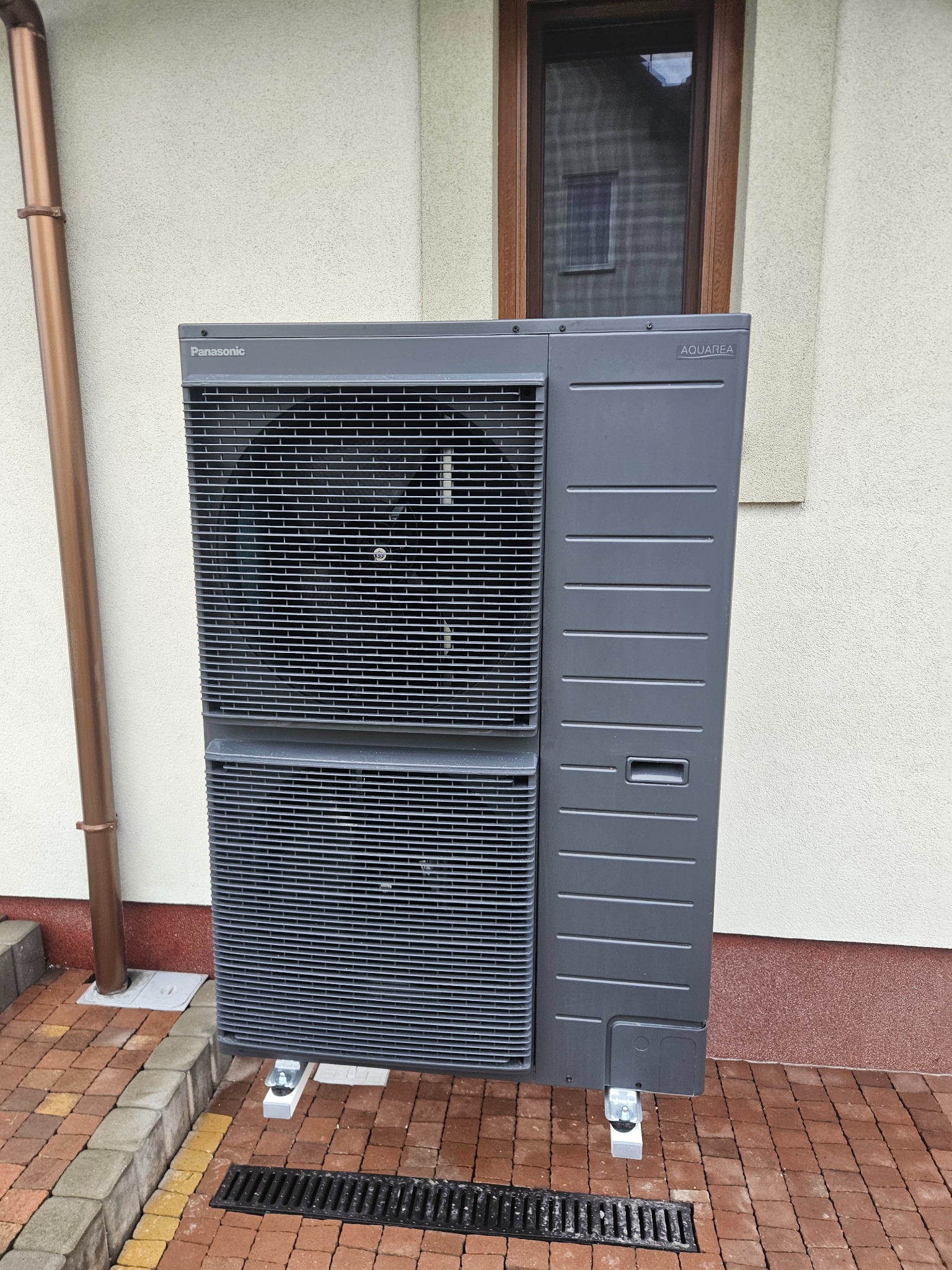 Szara pompa ciepła Panasonic Aquarea, zamontowana na zewnątrz budynku, widoczna z bliska na tle elewacji i kostki brukowej. Rynna odprowadzająca wodę po lewej stronie.