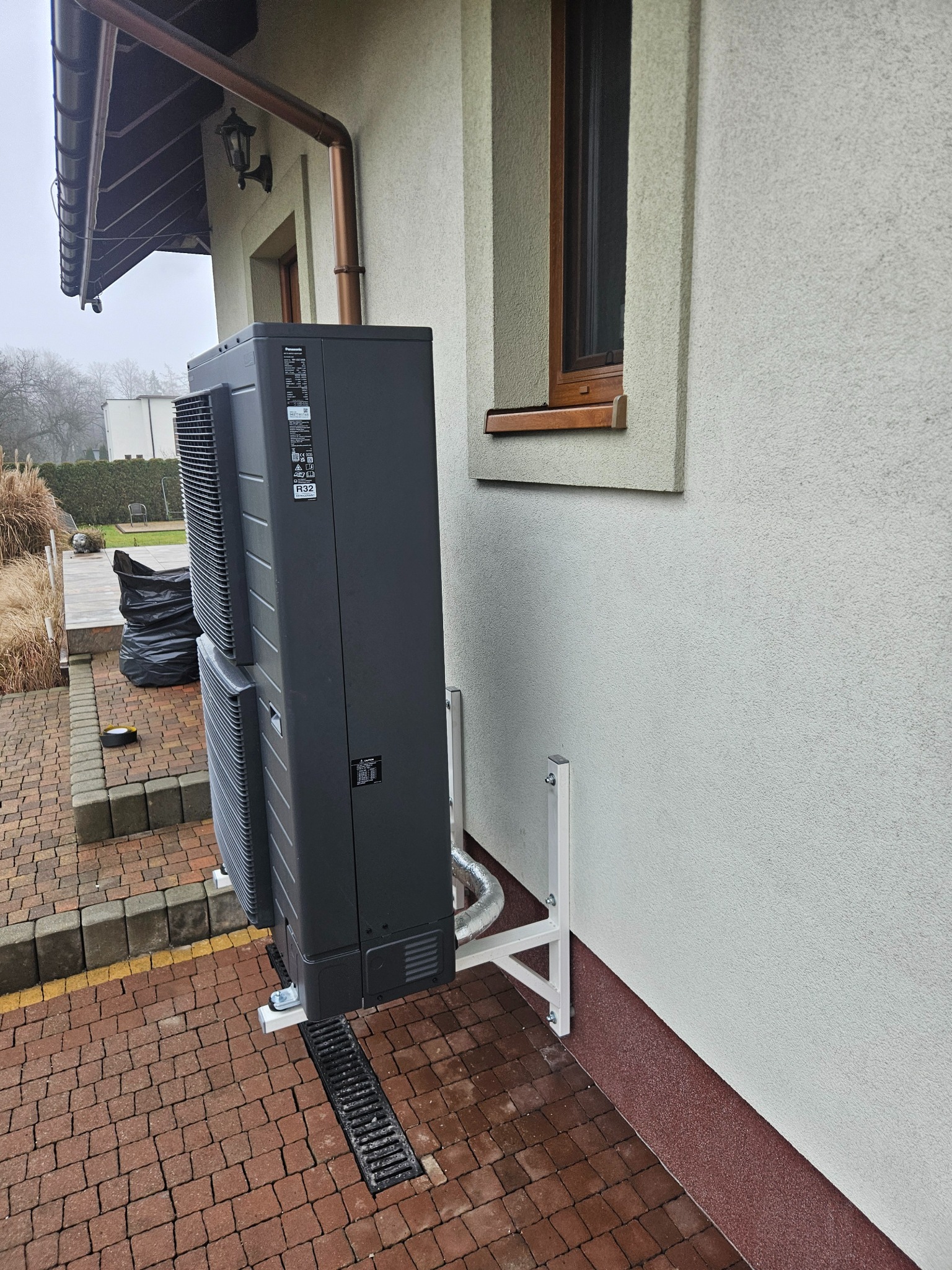 Zamontowana, czarna pompa ciepła Panasonic Aquarea na zewnątrz budynku, z widocznymi elementami instalacji i odpływem wody. Widok z boku, kostka brukowa.