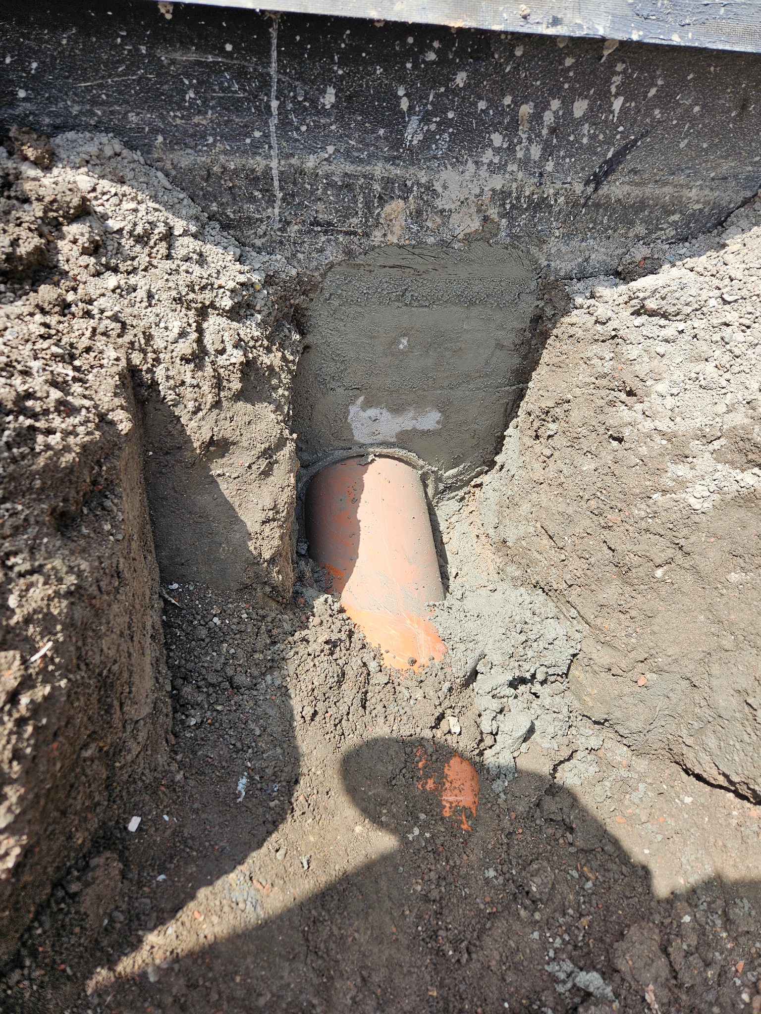Pomarańczowa rura kanalizacyjna w wykopie, częściowo obsypana ziemią i zabezpieczona betonem. Widoczne ślady prac ziemnych i cień osoby.