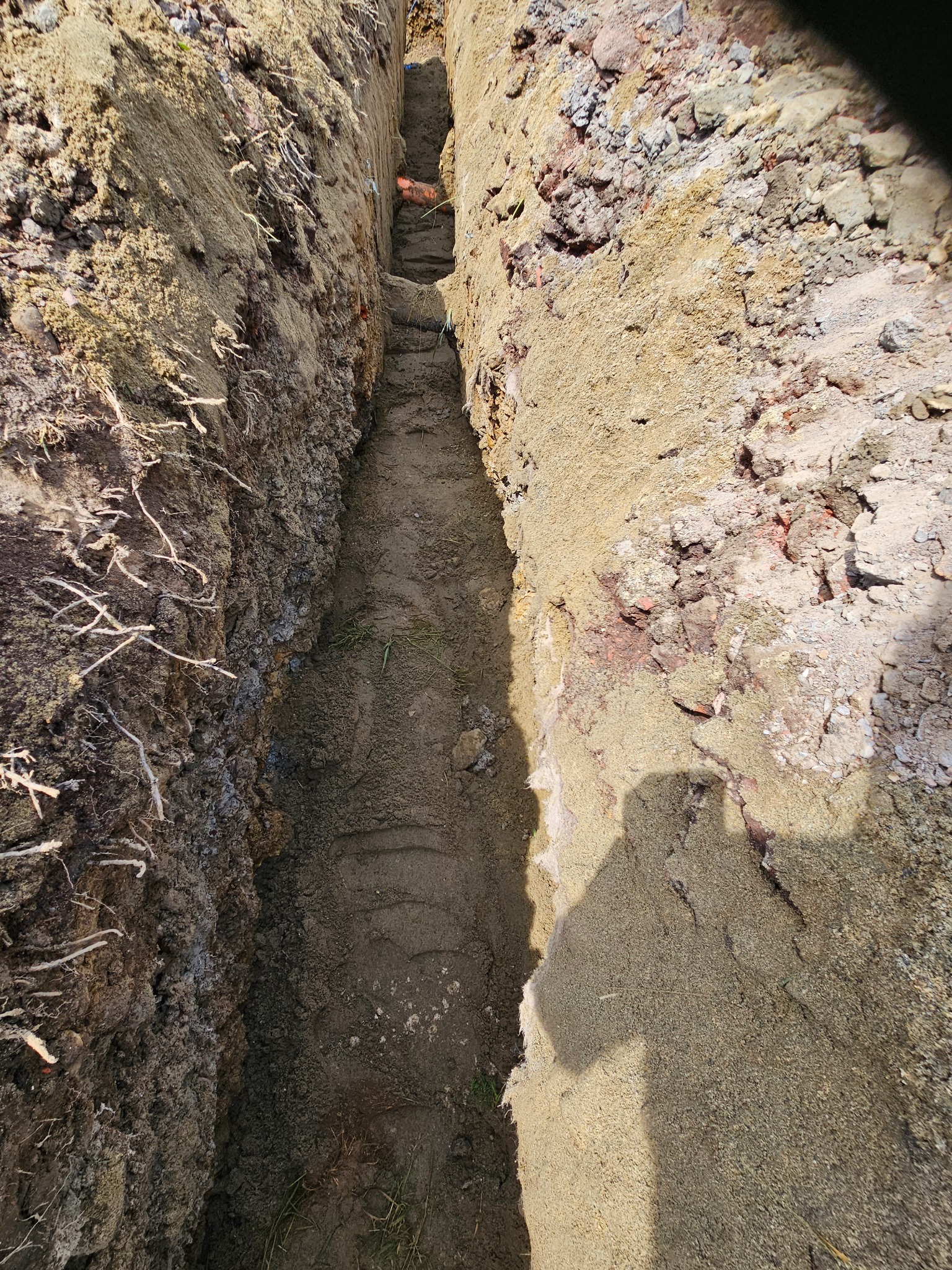 Wykopany wąski rów w ziemi, prawdopodobnie pod instalację. Widoczne ślady po pracy koparki i fragment pomarańczowej rury na dnie. Ujęcie z góry, w słoneczny dzień.