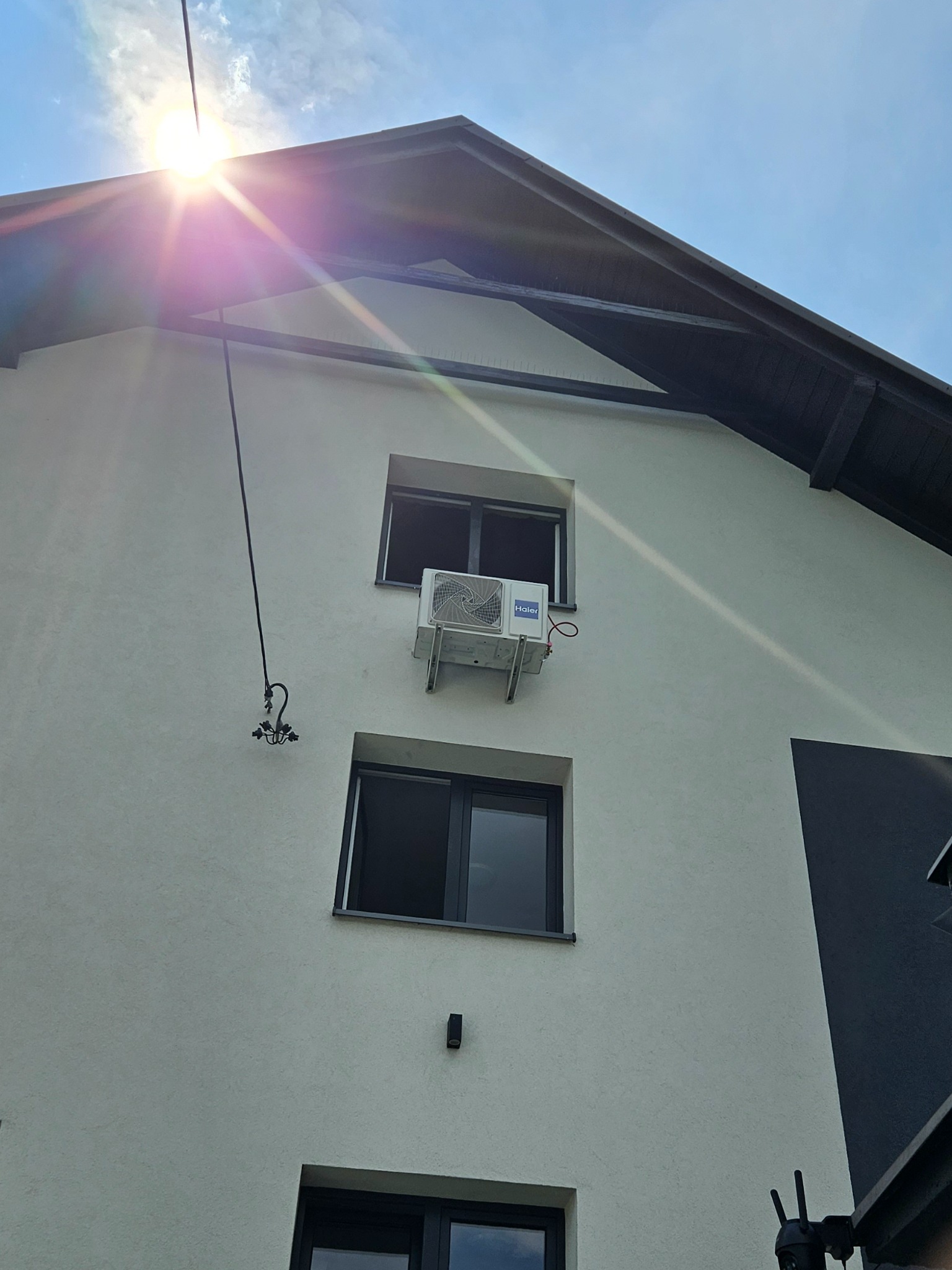 Biała elewacja domu z zamontowaną klimatyzacją Haier między oknami. Widoczne przewody i mocowanie. Słońce w górnym rogu kadru.