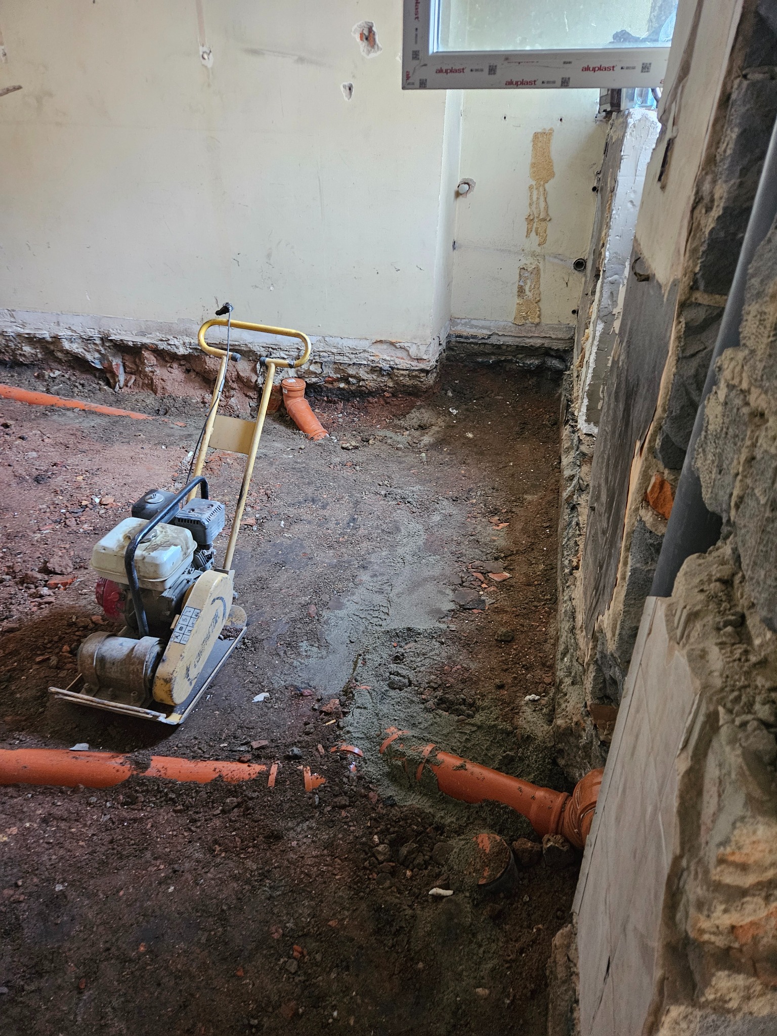 Wykop z pomarańczowymi rurami kanalizacyjnymi w trakcie układania, ubijarka w tle. Widoczne ślady po kuciu w ścianach. Przygotowanie podłoża pod instalację.