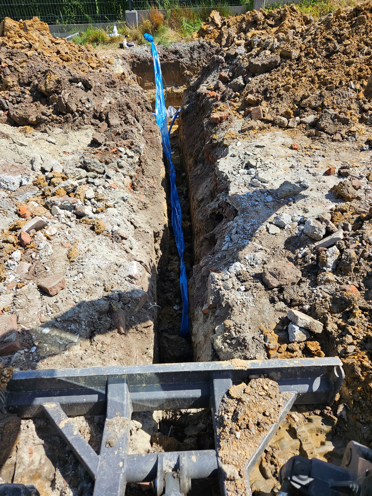 Wykopany wąski rów w ziemi, zabezpieczony niebieską folią. Widoczny fragment maszyny budowlanej. Grunt piaszczysto-gliniasty z gruzem. Etap prac ziemnych.