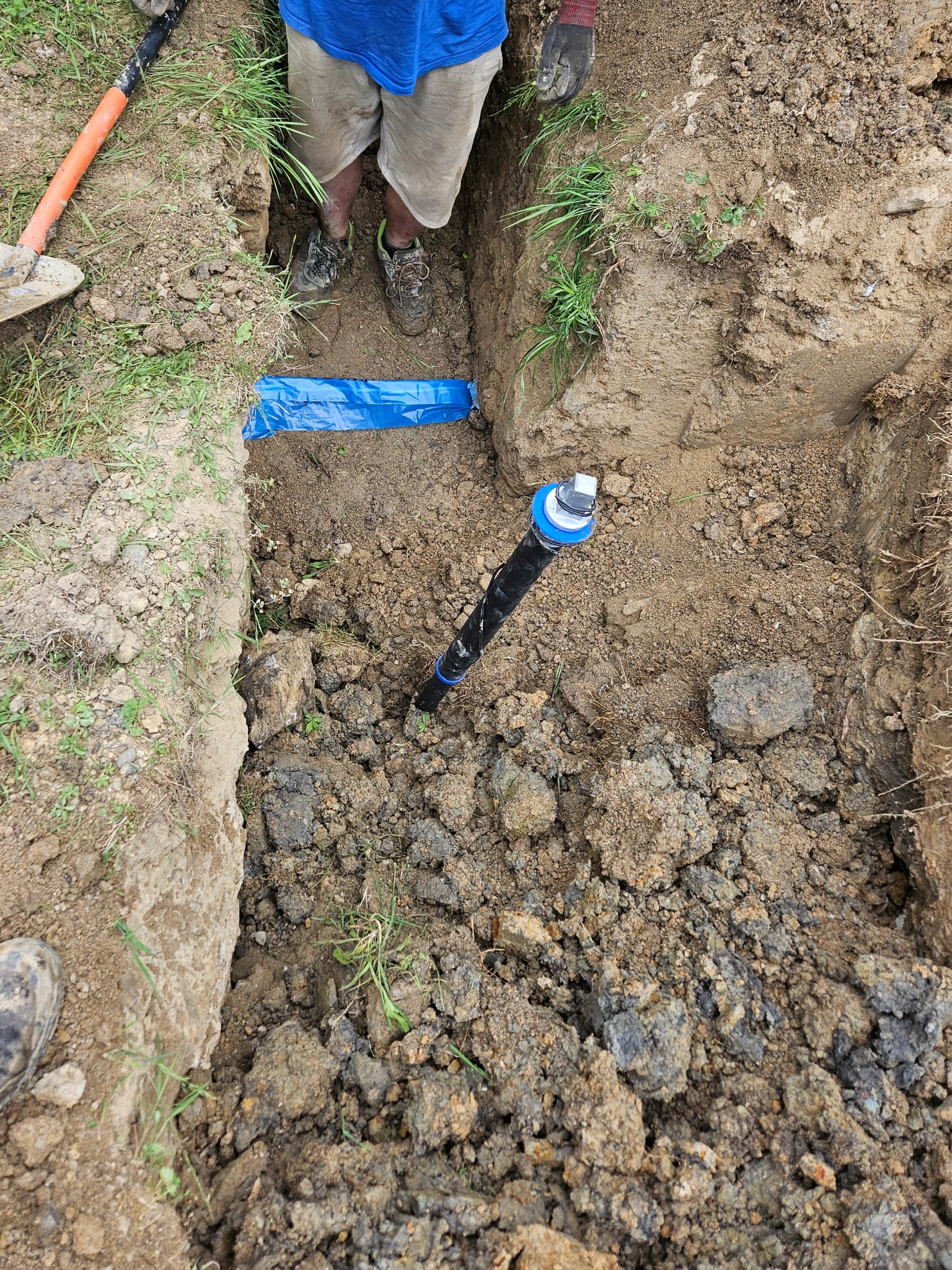 Wykop z rurą i niebieską folią w ziemi, widok z góry. Prace ziemne w Czechowice-Dziedzice. Widoczne narzędzia i fragment osoby.