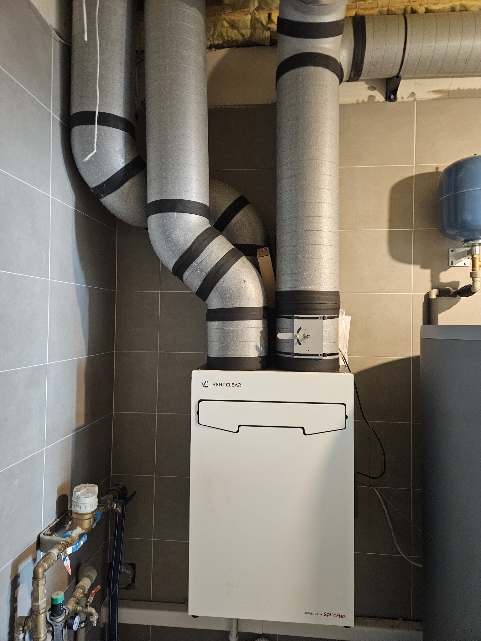 Biała centrala wentylacyjna Vent Clear z szarymi, izolowanymi przewodami wentylacyjnymi w pomieszczeniu wyłożonym szarą płytką. Widoczne przyłącze wod-kan.