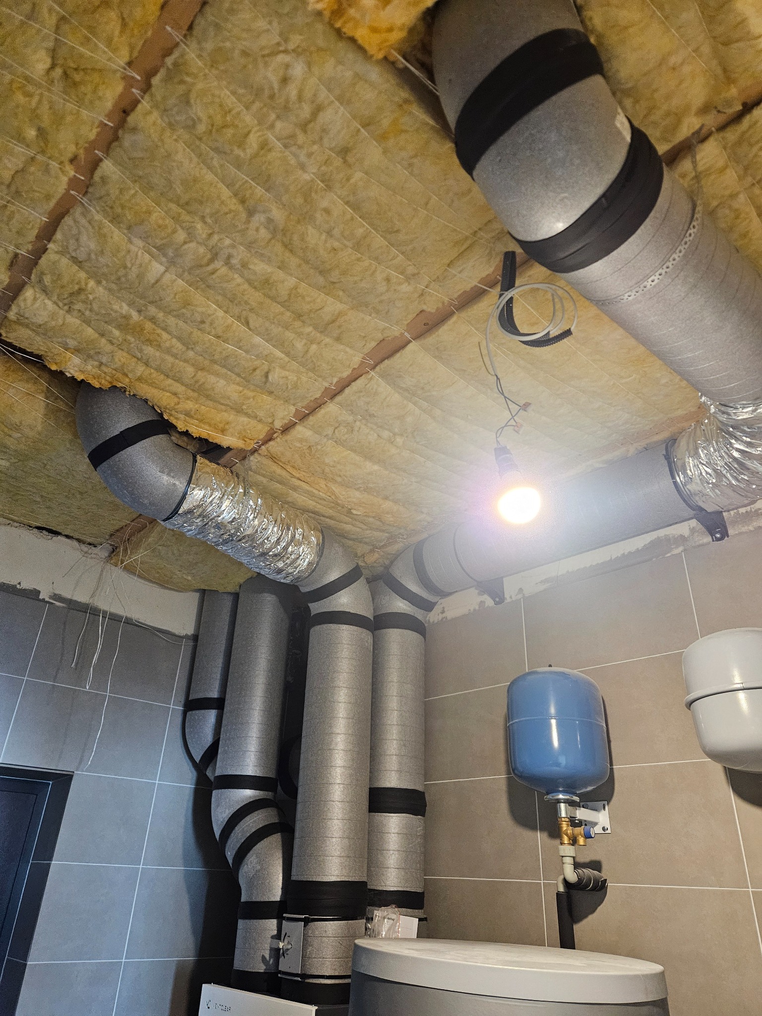 Instalacja wentylacyjna z izolacją na suficie, widoczne rury wentylacyjne z czarnymi opaskami, zbiornik wyrównawczy i oświetlenie żarówką. Szare płytki na ścianach.