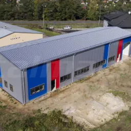Gotowa hala przemysłowa z szarej blachy trapezowej, z akcentami w kolorze niebieskim i czerwonym, widziana z lotu ptaka, na tle innych budynków i zieleni.
