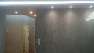 Ściana w odcieniach szarości z efektem dekoracyjnym, oświetlona punktowymi lampami sufitowymi i listwą LED, widok na korytarz z drzwiami.