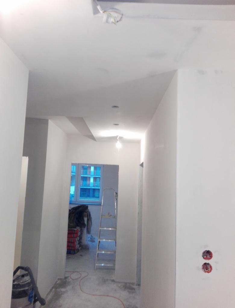 Korytarz w trakcie remontu: świeżo pomalowane ściany na biało, widoczne puszki elektryczne bez osprzętu, drabina aluminiowa, worki z zaprawą i odkurzacz przemysłowy.