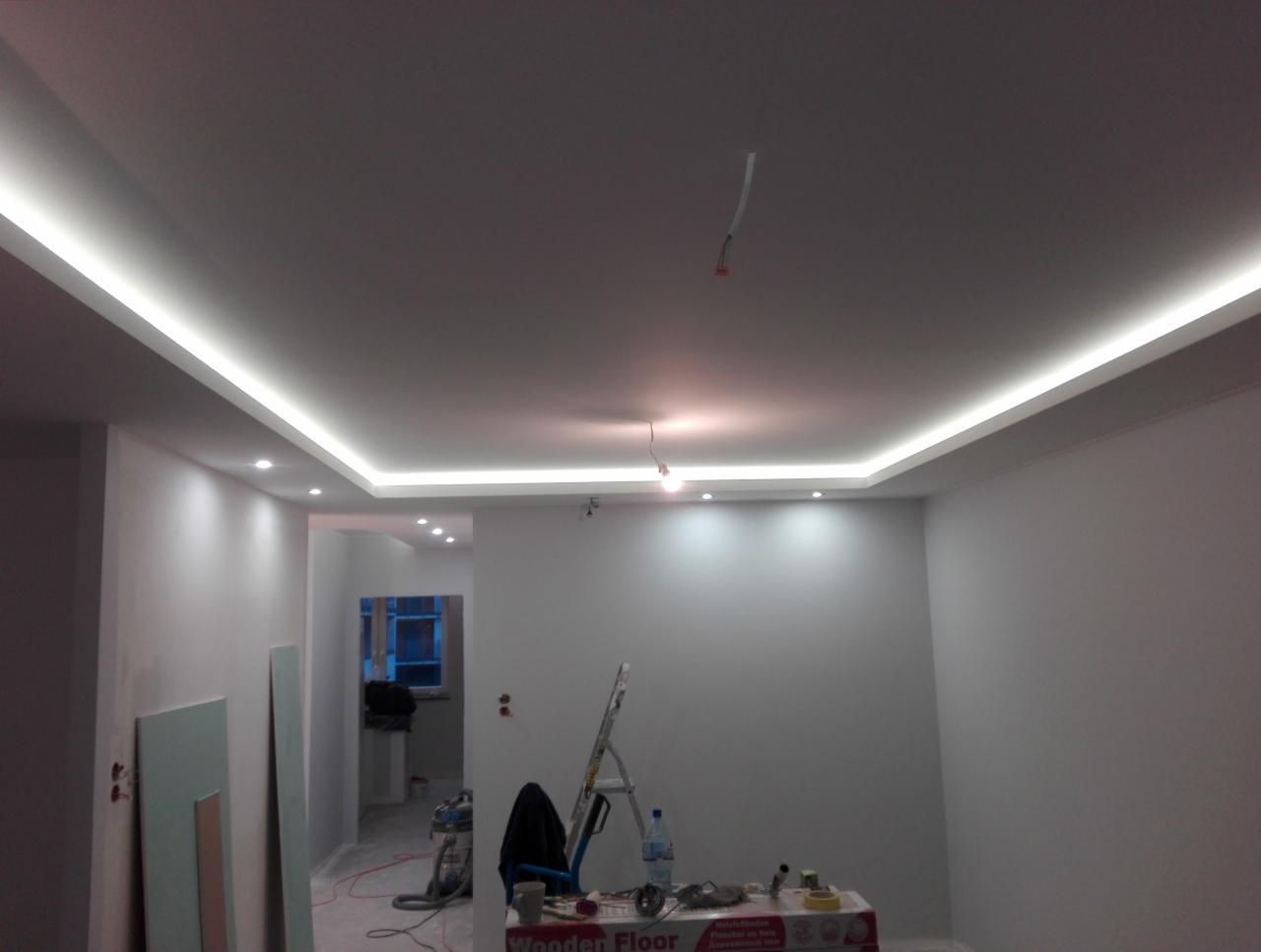 Wykończone wnętrze z podwieszanym sufitem i oświetleniem LED, widoczne narzędzia malarskie i drabina, ściany przygotowane do dalszych prac wykończeniowych.