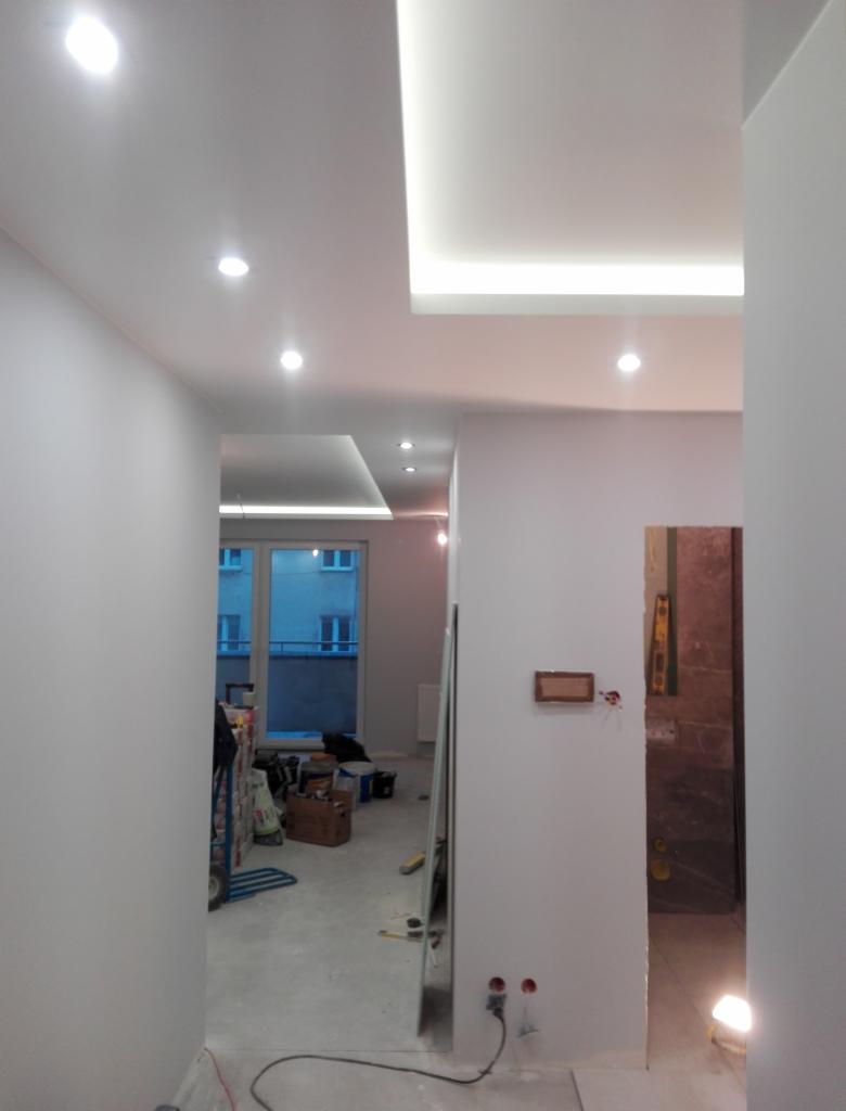 Wnętrze mieszkania w trakcie remontu, świeżo pomalowane ściany na biało, widoczne przygotowania do dalszych prac wykończeniowych, narzędzia i materiały budowlane w tle.