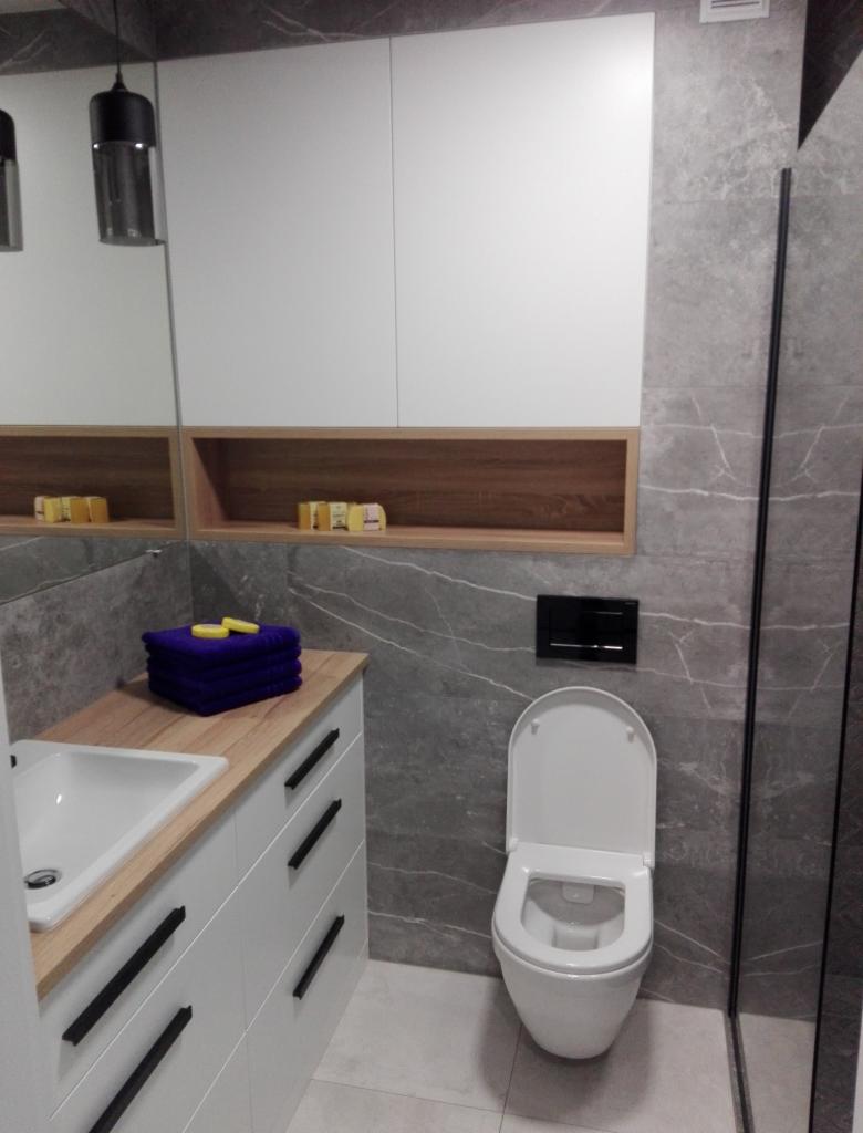 Nowoczesna łazienka z szarymi płytkami imitującymi marmur, białą szafką z umywalką i drewnianym blatem, wisząca toaleta z czarnym przyciskiem oraz czarna lampa wisząca.