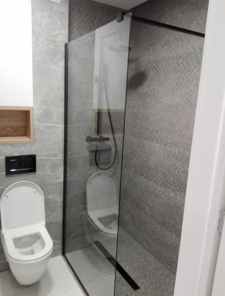 Nowoczesna łazienka z szarymi płytkami imitującymi jodełkę na ścianie prysznica, kabina prysznicowa typu walk-in z czarnymi profilami, wisząca toaleta z czarnym przyciskiem spłukującym.