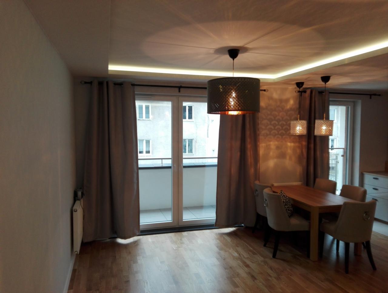 Jasne wnętrze jadalni z drewnianym stołem i krzesłami w kolorze beżu, oświetlone trzema wiszącymi lampami z ażurowymi kloszami i listwą LED pod sufitem, z widokiem na balkon przez duże okno przesuwne.