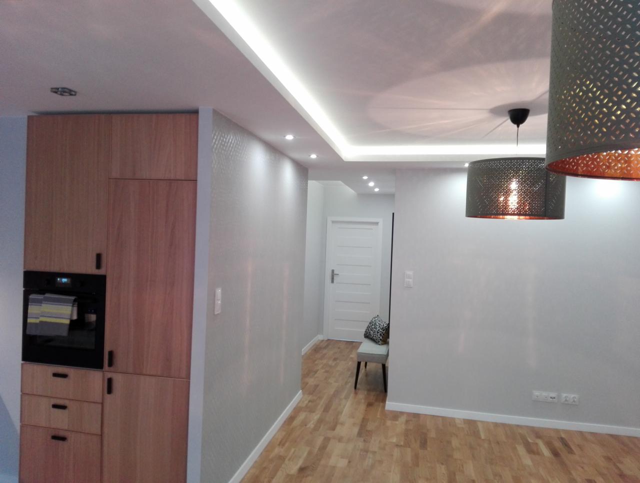 Jasne wnętrze w Gdańsku z drewnianą podłogą i szafkami, ściany wykończone w odcieniach szarości z delikatną fakturą, nowoczesne lampy wiszące z ażurowymi kloszami.