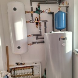 INSTAL-TOM TOMASZ KWAŚNIK - Instalacja grzewcza z dwoma zbiornikami, rurami i rozdzielaczem podłogówki. Widoczne elementy marki Ferroli i Uponor. Szare rury mocowane do ściany.