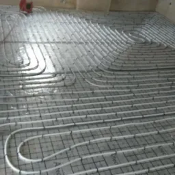 Białe rury ogrzewania podłogowego ułożone na siatce w nowym budynku, widoczne okno i elementy konstrukcyjne ścian.