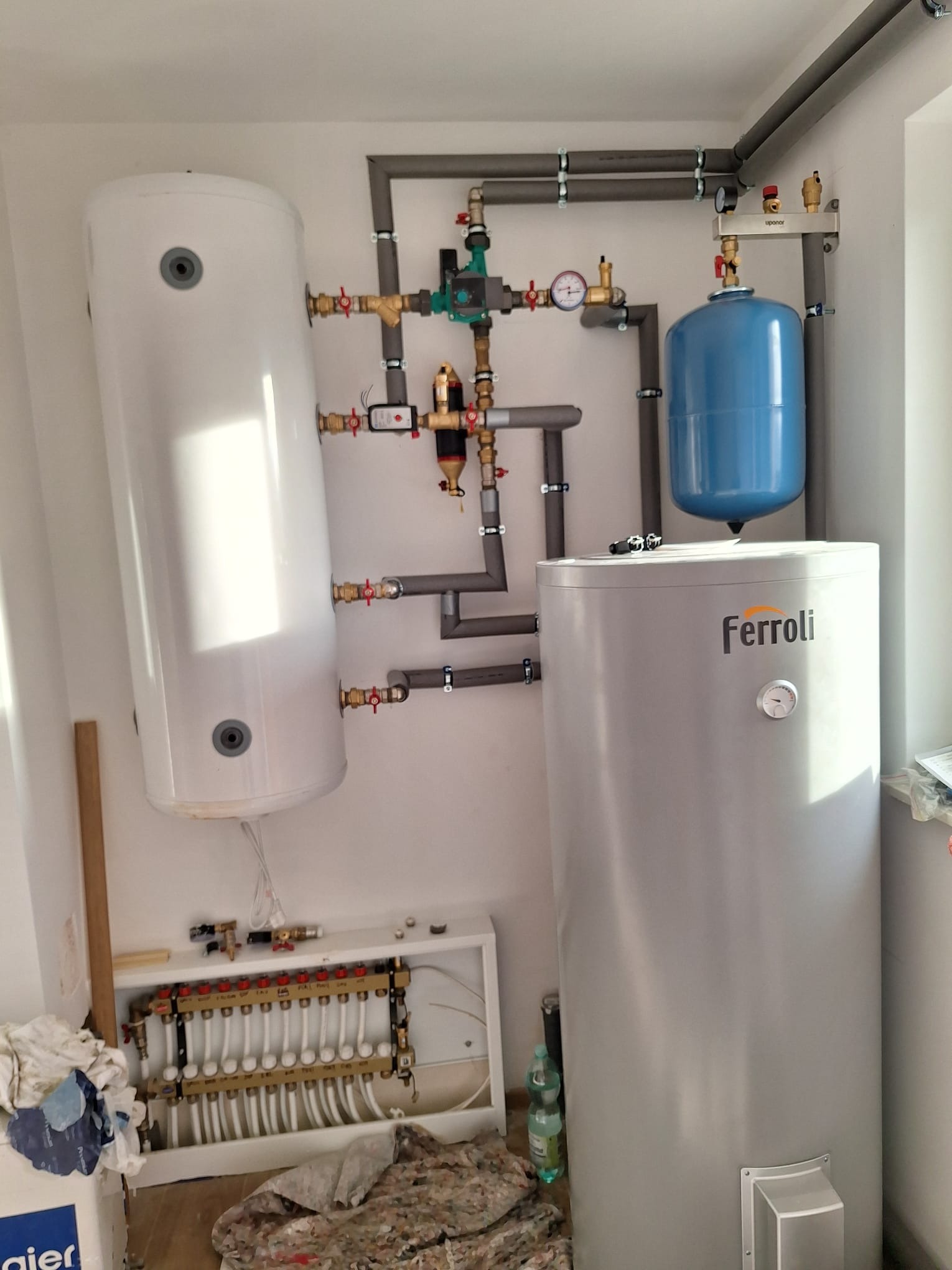 Instalacja grzewcza z dwoma zbiornikami, rurami i rozdzielaczem podłogówki. Widoczne elementy marki Ferroli i Uponor. Szare rury mocowane do ściany.