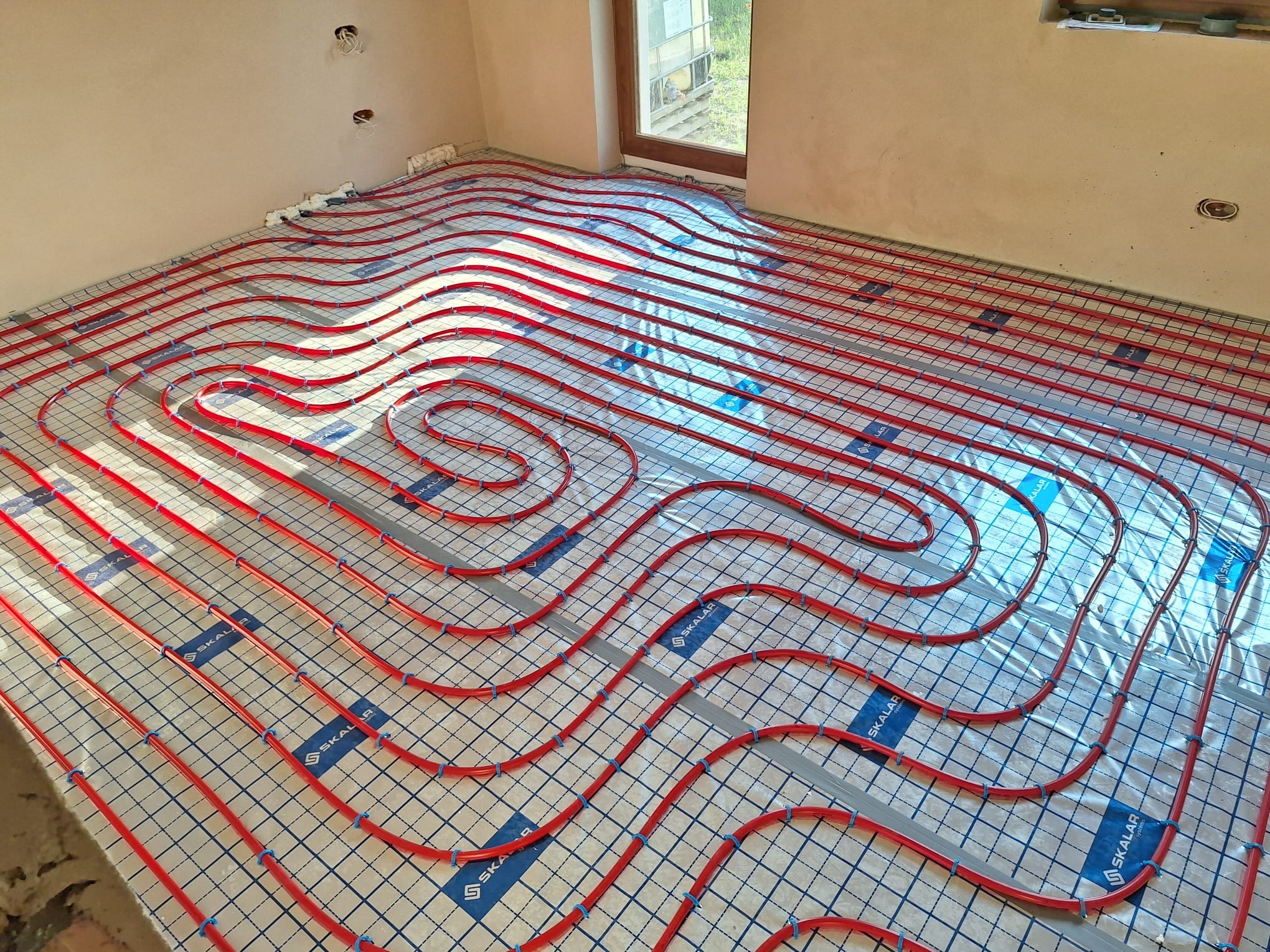 Czerwone rury ogrzewania podłogowego ułożone na siatce w jasnym pomieszczeniu z oknem. Widoczne niebieskie uchwyty mocujące i fragmenty pianki izolacyjnej przy ścianie.