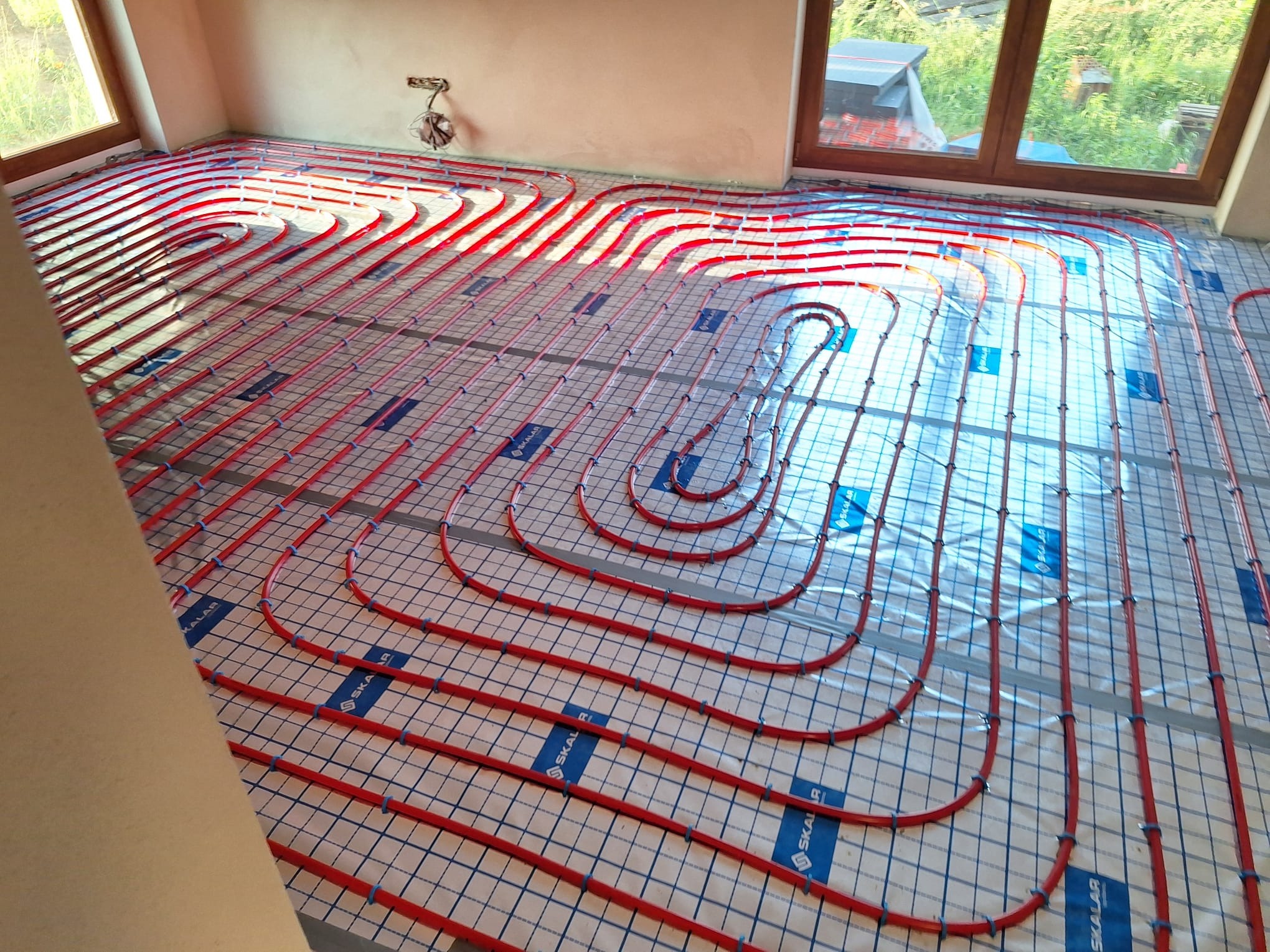 Czerwone rury ogrzewania podłogowego ułożone na folii z niebieską siatką w jasnym pomieszczeniu z oknami. Widoczne uchwyty mocujące rury.