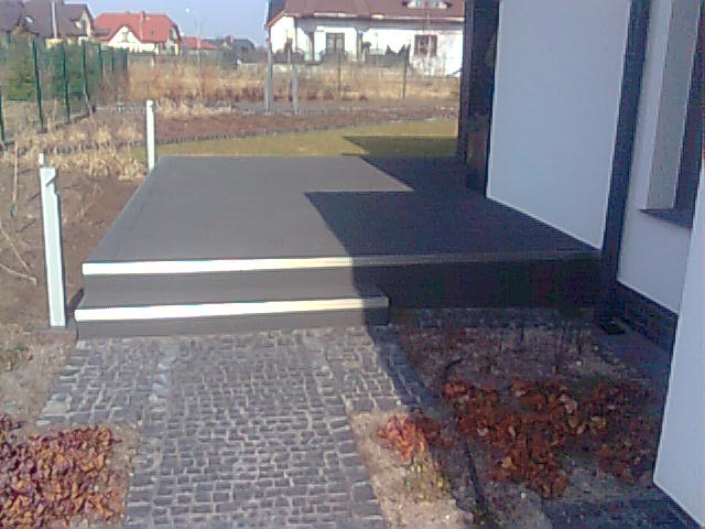 Nowoczesny taras z szarego kompozytu, z dwoma stopniami i białymi listwami wykończeniowymi, prowadzący do domu z białymi ścianami i kostką brukową na ziemi. W tle widoczne zabudowania.