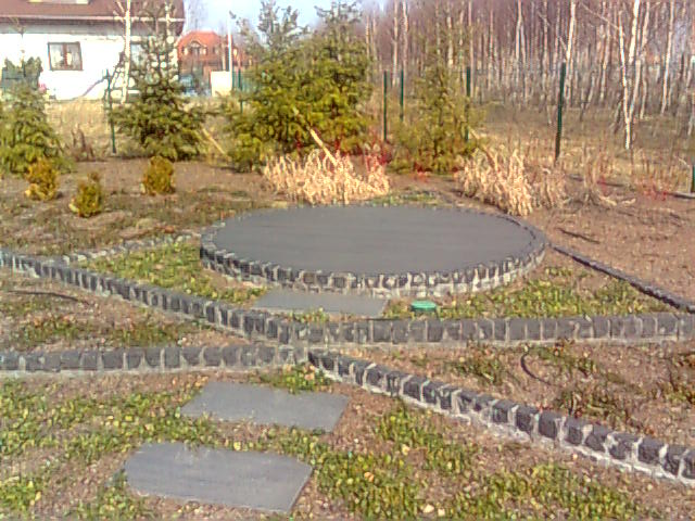 Ogród z okrągłym tarasem wyłożonym szarą nawierzchnią, obramowanym kostką brukową, z ścieżką z kamiennych płyt i obrzeżami z kostki, na tle drzew i domu.