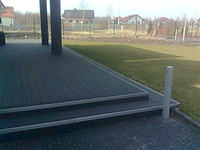 Zewnętrzny taras z ciemnych desek kompozytowych, dwustopniowe schody z aluminiowymi listwami, widok na trawnik i zabudowę jednorodzinną w tle.