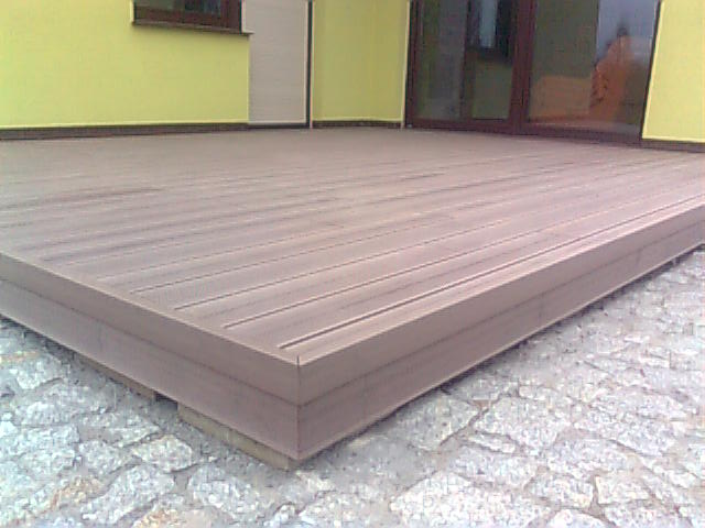 Nowo wykonany taras z desek kompozytowych na kamiennym podłożu, widok z niskiej perspektywy, ściana budynku w tle.