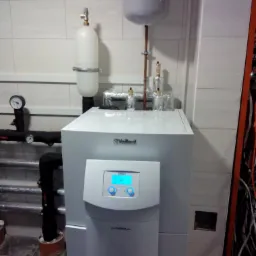 Biały piec grzewczy Vaillant z podłączonymi rurami i zbiornikami wyrównawczymi, zainstalowany w pomieszczeniu z białymi płytkami.