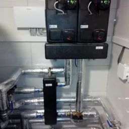 Zamontowana instalacja pompy ciepła z czarnymi modułami sterującymi z manometrami i rurami w izolacji aluminiowej, widoczny zbiornik wyrównawczy.