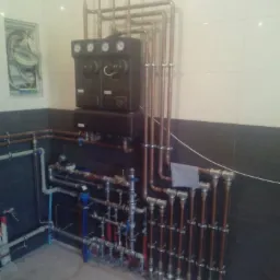 Złożona instalacja hydrauliczna w kotłowni, z licznymi rurami miedzianymi i zaworami, manometry na górnym panelu urządzenia, widoczne połączenia i mocowania do ściany wyłożonej płytkami ceramicznymi.