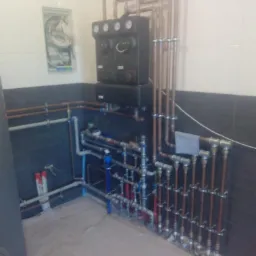 Złożona instalacja z rur miedzianych i plastikowych, z czarnym panelem sterującym z manometrami, zamontowana w pomieszczeniu z białymi i ciemnymi płytkami ceramicznymi.