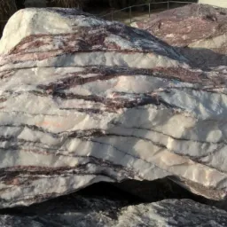 Duży, nieregularny kamień dekoracyjny z białymi i bordowymi pasmami, ułożony na innych kamieniach w słoneczny dzień.