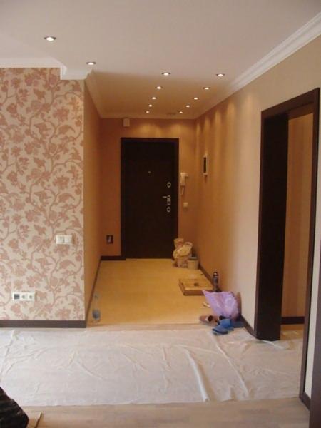 Korytarz w trakcie remontu: ściana z tapetą w kwiaty, pomalowane na beżowo ściany, zabezpieczona folią podłoga i widoczne narzędzia malarskie.