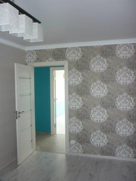 Pokój po tapetowaniu z wzorzystą tapetą w odcieniach szarości i bieli, widoczna otwarta, biała, panelowa drzwi do pomieszczenia z turkusową ścianą.