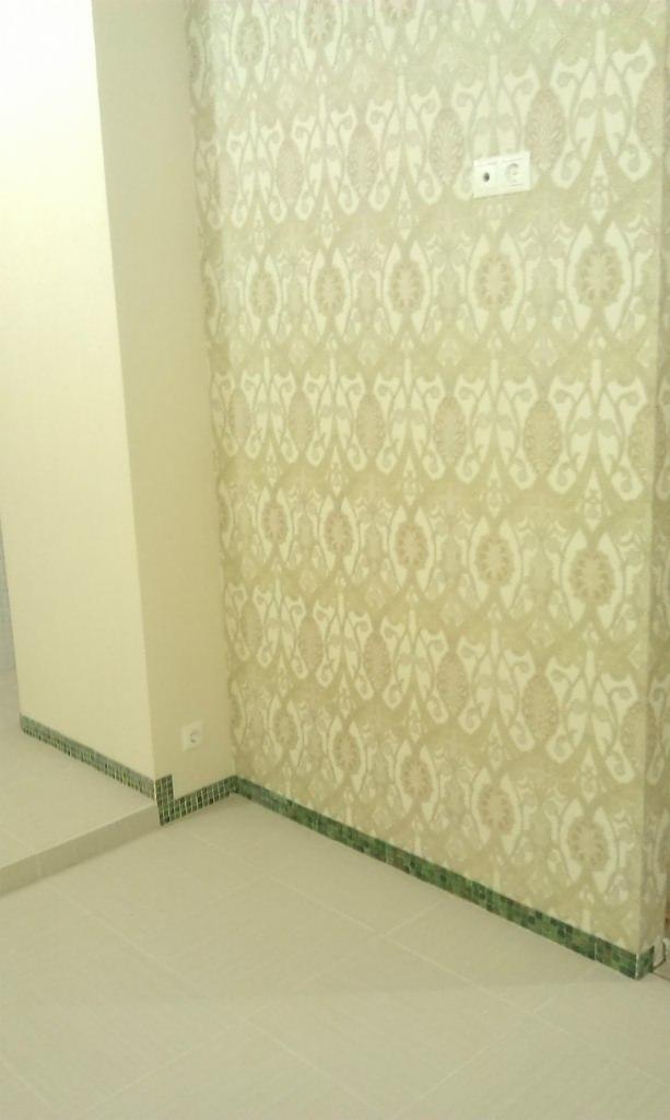 Wnętrze pokoju z tapetą w odcieniach beżu z ornamentacyjnym wzorem, wykończone zielonym cokolikiem z mozaiki, gładkie, kremowe ściany i jasna, kwadratowa podłoga z płytek ceramicznych.