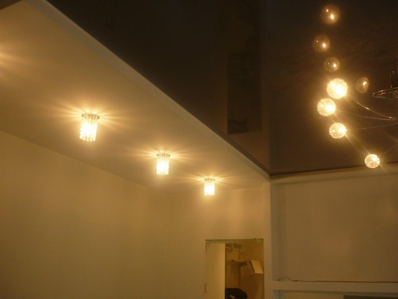 Błyszczący, ciemny sufit napinany z trzema kryształowymi lampami punktowymi wbudowanymi w biały sufit podwieszany oraz wiszący żyrandol z kulistymi kloszami.