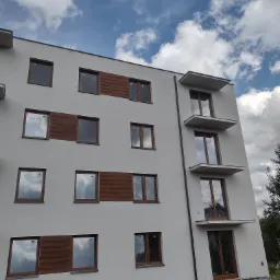 Nowoczesny budynek wielorodzinny z szarą elewacją, drewnianymi panelami dekoracyjnymi i balkonami zadaszonymi, pod błękitnym niebem z białymi chmurami.