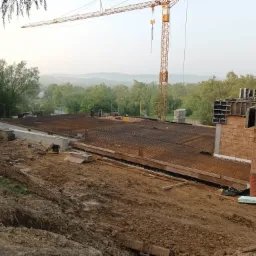 Zbrojenie płyty fundamentowej domu w trakcie budowy, widoczny żuraw budowlany, teren wokół w stanie surowym, w tle zalesione wzgórza.