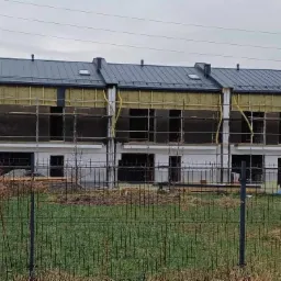 Szeregówka w trakcie prac elewacyjnych: widoczne rusztowania, izolacja termiczna na fragmentach ścian oraz szara blacha na dachu w pochmurny dzień.